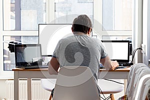 A man in a gray t-shirt is sitting at a workplace with two laptops and a monitor near the window. Remote work from home photo