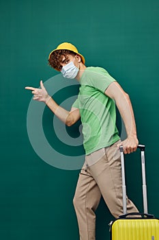 man on a green background, dressed in a yellow panama hat and with a mask on his face, holding a bright travel suitcase