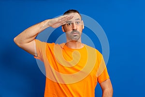 A man in greeting pose and serious face. A hispanic man with a beard wearing orange t-shirt isolated on blue background