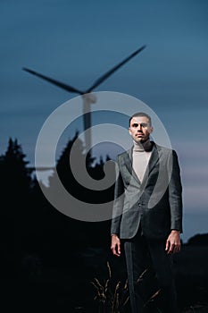 A man in a grey business suit stands next to a windmill after sunset .Businessman near windmills at night.Modern concept of the