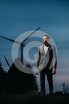 A man in a grey business suit stands next to a windmill after sunset .Businessman near windmills at night.Modern concept of the