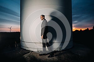 A man in a grey business suit stands next to a windmill after sunset .Businessman near windmills at night.Modern concept of the
