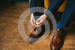 A man or a groom in a blue suit ties up shoelaces on brown leather shoes brogues on a wooden parquet background photo