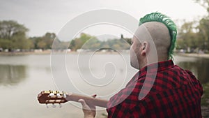 A man with a grunge style playing an acoustic guitar beside lake