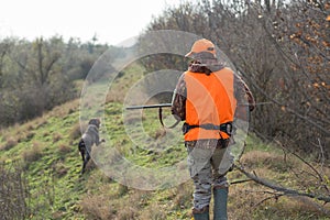 A man with a gun in his hands and an orange vest on a pheasant hunt in a wooded area in cloudy weather.