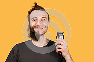 Man with half shave beard surprised with wide eyes hold hair clipper. Isolated on orange background