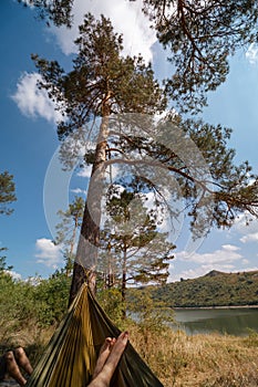 Man in hammock, first person look view, warm summer day, pine tree. river and mountains background. Travel and vacation, tourism