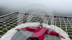 Man on hammock net lying and resting looking into stormy sky in the mountains. High above cloudy abyss. Thick white fog