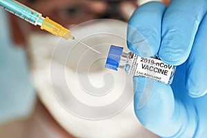 Man hand in blue nitrile glove hold small vial with coronavirus vaccine, to inject it with green orange syringe, blurred face