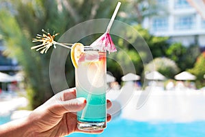 Man hand with coloured coctail with piece of orange lemon on hotel tropical background, cold fresh drink cocktail on palm tropical