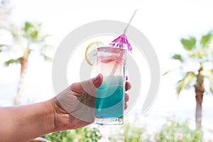 Man hand with coloured coctail with piece of orange lemon on hotel tropical background, cold fresh drink cocktail on palm tropical