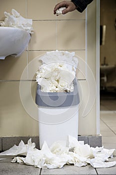 Man hand drops paper tissue in full trash bin, utilized toilet paper dropped around the trash basket. Dirty toilet