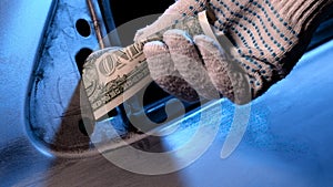 A man hand in a glove holds one dollar bill instead of a mirror on the car door. A scratched metal surface is prepared for