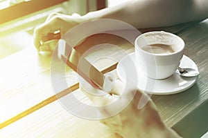 man hand holding a cell phone and drink coffee on a wooden table in a coffee shop