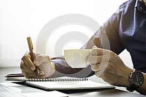 man hand holding coffee cup with writing pen memo into notebook paper on table desk office.