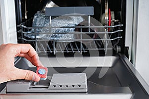 A man hand holding dishwasher detergent tablet. Putting tab into dishwasher, close up.  Dishwasher machine full loaded