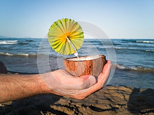 Man hand holding a halved coconut with a small green umbrella in the beach