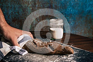 A man hand holding a handmade rye bread wrapped with a cloth over a stone table and a flour jar next to it