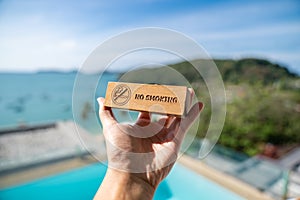 Man hand holding no smoking sign written on wood panel with beach background