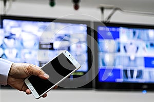 A man hand holding a smart phone against blurred CCTV security camera monitor in a building.