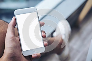 A man hand holding white mobile phone with blank screen on thigh with white canvas shoes in modern cafe with feeling relax