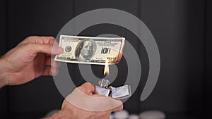 A man hand holds 100 us dollars in his hands and set fire to a lighter from below. A dollar bill burns with flames on a