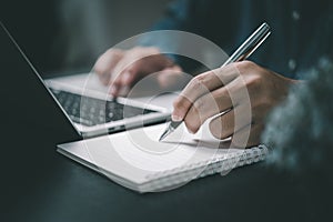 Man hand with pen writing on notebook with computer laptop at desk