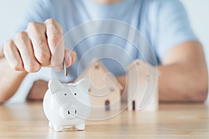 Man hand put coin into White piggy bank with wooden house on wood table. saving money for buying house, financial plan home loan