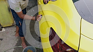 Man hand washing a yellow sports car before painting with a sponge and bucket of water focusing on the rear side