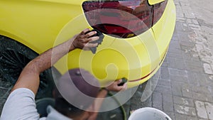 Man hand washing a yellow sports car before painting with a sponge and bucket of water focusing on the rear side