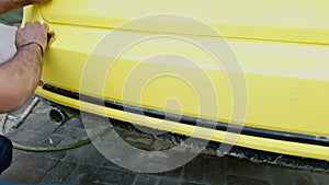 Man hand washing a yellow sports car before painting with a sponge and bucket of water focusing on the rear side