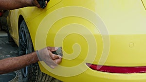 Man hand washing a yellow sports car before painting with a sponge and bucket of water focusing on the rear side