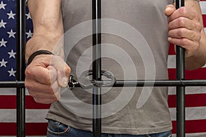 A man is handcuffed in a cell behind bars against the background of an American flag. Concept: the prisoner in the courtroom, the