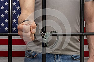 A man is handcuffed in a cell behind bars against the background of an American flag. Concept: the prisoner in the courtroom, the