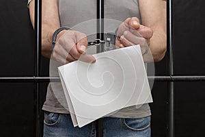 A man in handcuffs holds a white sheet of paper in his hands behind bars in a cell. Concept: a petition for clemency, the last wor