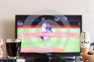 Man hands holding cup of cola and ice cream cone with a TV broadcasting baseball match on background