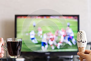 Man hands holding cup of cola and ice cream cone with a TV broadcasting football match on background