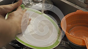 A man hands wash a plate in a metal sink with a pile of dishes in a close-up
