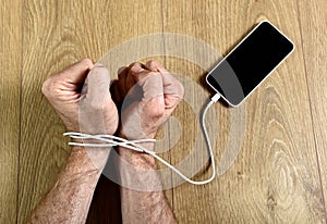 Man hands wrapped on wrists with mobile phone cable handcuffed in smart phone networking addiction concept