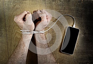 Man hands wrapped on wrists with mobile phone cable handcuffed in smart phone networking addiction concept