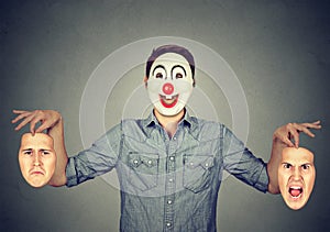 Man in happy clown mask holding two faces expressing anger and sadness