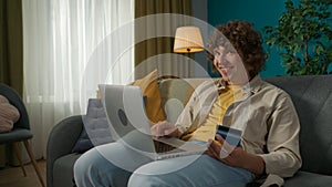 Man with a happy smile sitting on the sofa in the living room and holding a credit card and a laptop, online shopping