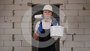 A man in a hard hat stands on a construction site with paint and a roller.