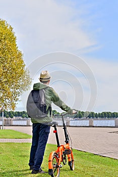 Man in hat and with backpack, standing with a bicycle in city park.