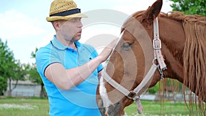 The man in hat caress the horse by the muzzle on meadow