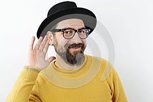 Man with hat and eyeglasses smiling with hand over ear listening and hearing to rumors or gossip.