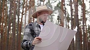 Man in a hat holds a map while hiking in the forest. Orientation concept,
