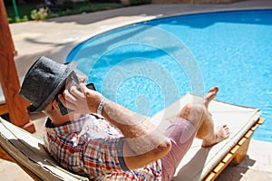 Man in a hat lying on a lounger by the pool and talking on the phone
