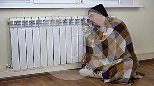 A man in a hat and a plaid at the radiator on the floor. A young man is freezing at home because of non-payment of