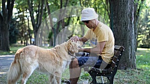 Man in a hat plays with his pet dog in the park. Golden retriever. Man with his dog are sitting on the bench in the park